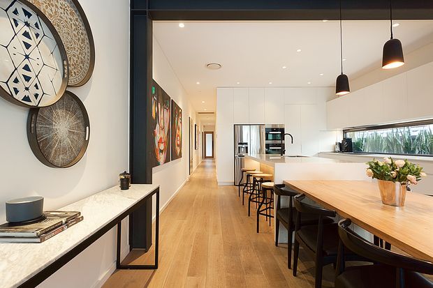 A bright, open-plan contemporary kitchen and dining area with a long light-wood dining table, black chairs, white flat-panel cabinetry, integrated stainless-steel appliances, and a pale stone island benchtop. Black pendant lights hang above the dining table, while a slim console with decorative wall discs and artwork lines the adjacent wall. Timber flooring extends through the kitchen into a hallway, creating a warm, spacious interior.