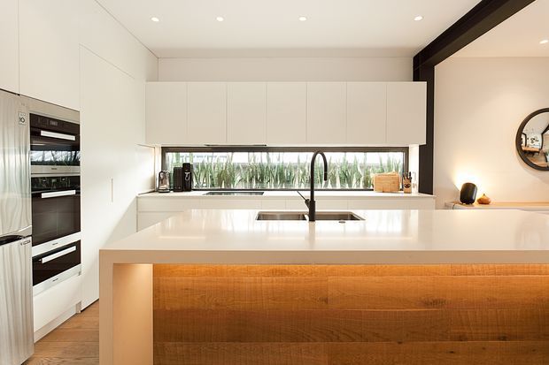 Bright contemporary kitchen viewed from the living area, centred on a large white island with integrated sink and curved black tap. The kitchen features extensive white handleless cabinetry, built-in appliances, a long horizontal window with greenery outside, a white benchtop and a warm timber-clad island front. Recessed ceiling downlights and a dark-framed opening add architectural contrast.