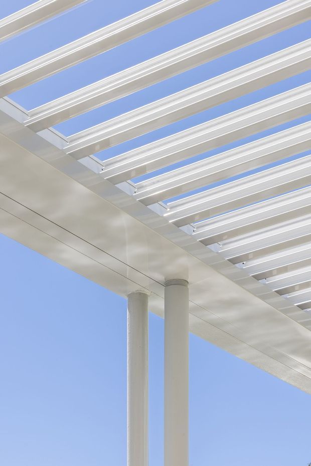 Close-up exterior architectural view of a white pergola or louvred canopy, with repeated linear roof beams supported by slender round columns against a clear blue sky. The image emphasizes clean geometric structure, open-air shading, and modern minimal detailing.