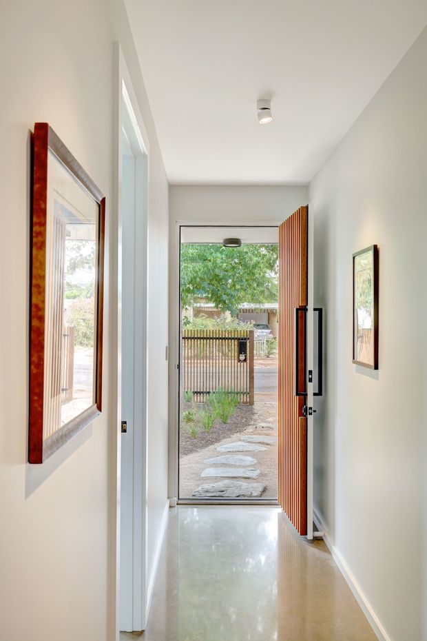 A narrow, bright residential entry hall viewed from inside toward an open front door. The hallway has smooth white walls and ceiling, a polished pale concrete or stone-like floor, recessed or surface-mounted ceiling lighting, framed artwork, and a striking vertically slatted timber front door. Through the doorway, a small landscaped path and timber fence are visible outside.