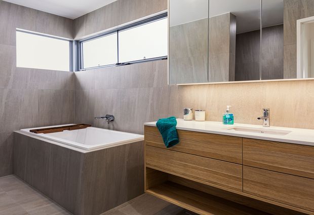 Contemporary bathroom with a built-in freestanding-style bathtub, wide horizontal windows, large-format grey-brown wall and floor tiles, and a long timber-look floating vanity with integrated basin. Mirrored cabinetry and warm under-cabinet lighting create a clean, refined finish.