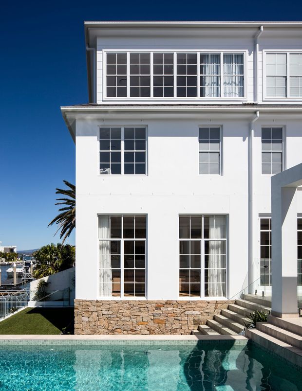 A bright exterior view of a multi-storey white rendered house with a strong classical influence, extensive multi-pane windows, projecting eaves, and a natural stacked-stone base. A swimming pool occupies the foreground, with steps leading to a glazed entry or terrace area. Palm trees and neighbouring buildings are visible in the background under a clear blue sky.