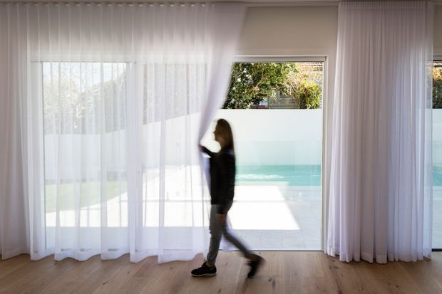 Bright contemporary interior with full-height sheer curtains framing expansive glazed doors or windows, opening to a swimming pool outside. A person walks across a light timber floor, while the neutral walls and abundant daylight create an airy, minimal atmosphere.