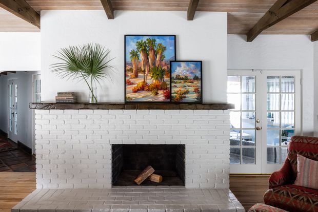 Bright living room centered on a white-painted brick fireplace with a broad hearth and dark timber mantel. Exposed timber ceiling beams and wood boards create a rustic architectural character, while French doors open to an adjacent space. Framed artwork, a potted palm, and a patterned upholstered armchair add warmth and colour.