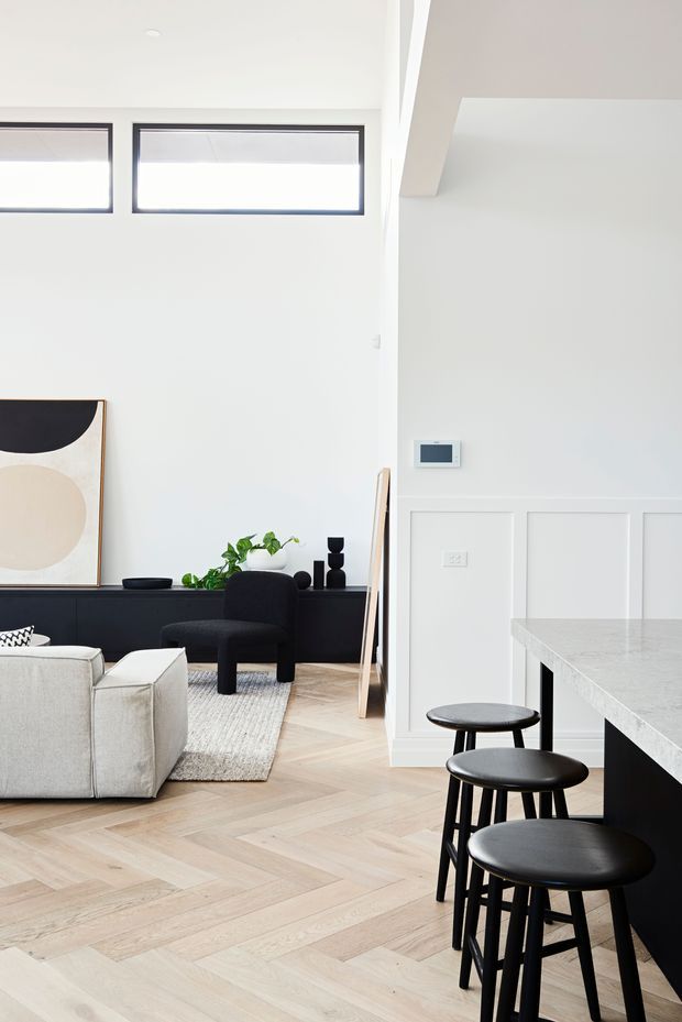 Bright open-plan interior combining a living room and kitchen. The room features pale herringbone timber flooring, white wall panelling, high clerestory windows with dark frames, a black built-in media console, neutral upholstered seating, sculptural artwork, and a dark kitchen island with a light stone benchtop and black stools. The overall look is clean, refined, and contemporary with strong black-and-white contrast.