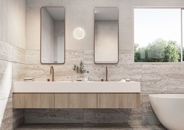 A refined contemporary bathroom featuring a long double vanity with light wood cabinetry, a white integrated benchtop, twin rounded-rectangle mirrors, and two warm metallic tap fixtures. Large beige-grey stone-look wall and floor tiles create a continuous finish, while a freestanding white bathtub and garden-facing window are visible on the right. The space has a calm, warm, natural material palette and soft daylight.
