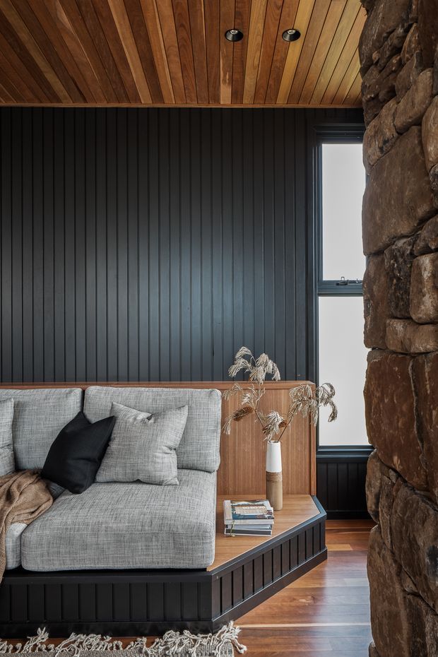 A moody, textural living room corner featuring a grey sectional sofa with black and grey cushions on a raised built-in platform. The room has dark vertical wall panelling, a warm timber-lined ceiling with recessed downlights, timber flooring, and a substantial rough-stacked natural stone wall. A timber built-in sideboard and angular shelf hold books and a vase with dried foliage, while a narrow window brings in natural light.