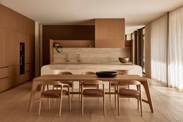 A warm, refined open-plan kitchen and dining area with extensive timber cabinetry, a stone kitchen island and backsplash, and a long timber dining table with matching upholstered chairs. Full-height sheer curtains line the right side, filtering soft natural light across the timber floor. The kitchen includes integrated appliances, a sculptural island, open shelving with greenery, and a central overhead timber volume.