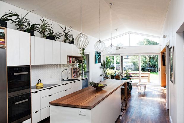 Bright contemporary open-plan kitchen and dining space with extensive white built-in cabinetry, integrated black appliances, a long timber island with bar seating, patterned tile splashback, pendant lights, indoor plants, and timber flooring. Glazed doors and a high triangular clerestory-style window bring in abundant natural light and connect the room to an outdoor area.