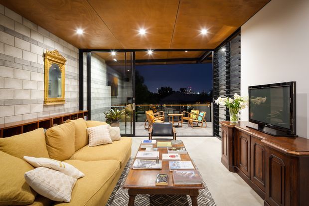 A warm, open-plan living room with a mustard-yellow sofa, patterned rug, timber coffee table, and traditional timber media cabinet. The room features exposed pale concrete block walls, a rich timber-panel ceiling with recessed spotlights, and extensive black-framed glazing opening to an outdoor terrace with seating and nighttime views. A gold-framed mirror and indoor plants add decorative detail.