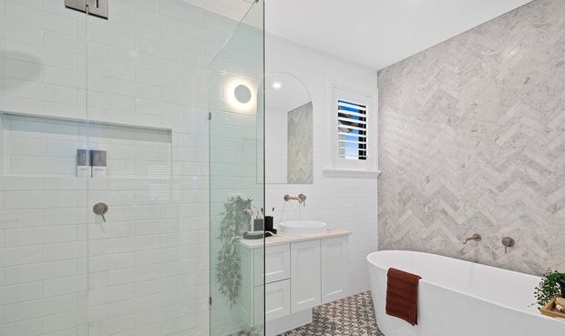 Bright contemporary bathroom featuring a frameless glass walk-in shower with white subway wall tiles, a wall-mounted white vanity, freestanding bathtub, patterned floor tiles, and a pale herringbone feature wall. A small shuttered window and mirror add light and reflection.