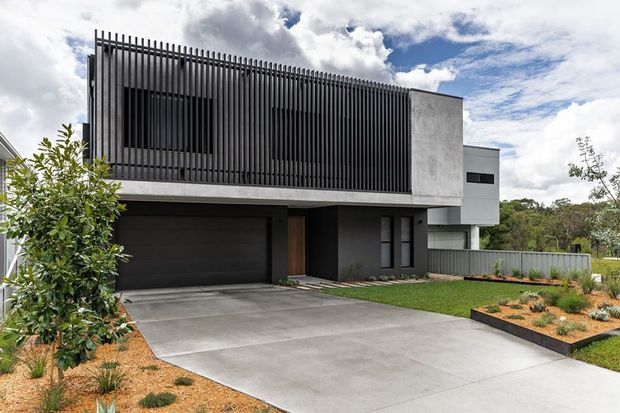 Contemporary two-storey residence viewed from the front, featuring a dark double garage, broad concrete driveway, strong horizontal and vertical facade elements, a tall black vertical-slat screen across the upper level, white rendered walls, and a landscaped front garden with low planting.