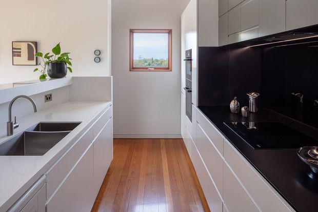A narrow, modern galley kitchen with white handleless cabinetry on both sides, a large dark stone or composite benchtop and splashback, integrated appliances, a double undermount sink, timber flooring, and a small timber-framed window at the end. The space is clean, compact, and strongly linear, with black and white finishes contrasted by warm wood.
