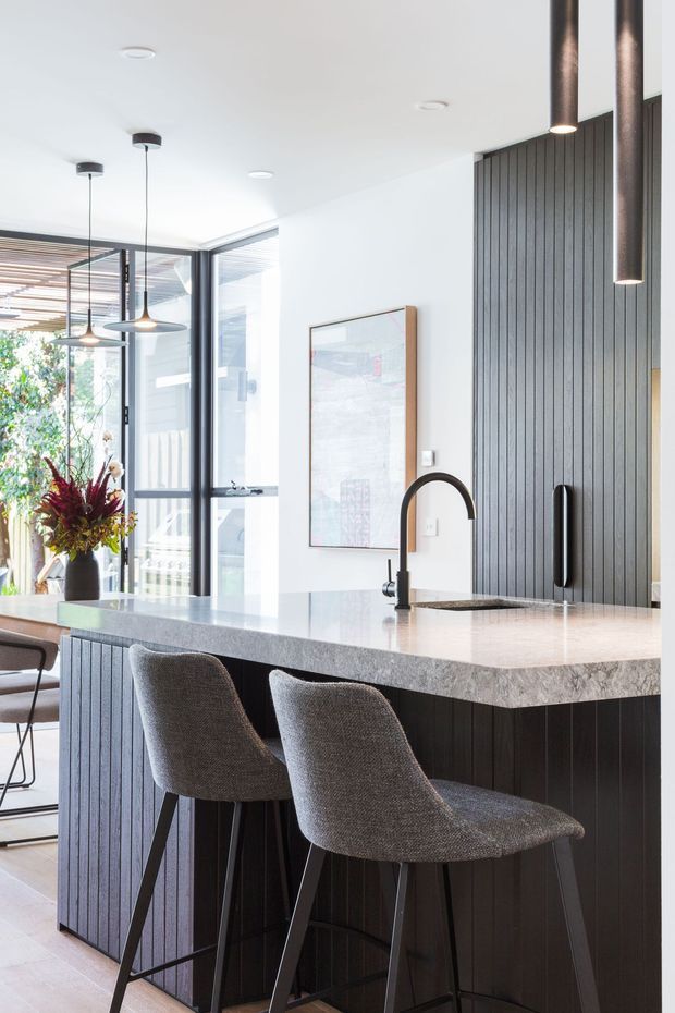 Bright contemporary kitchen interior featuring a large island with pale stone benchtop, dark vertically fluted cabinetry, integrated sink and black gooseneck tap, and two upholstered bar stools. Floor-to-ceiling glazing opens to a leafy outdoor area, while pendant and cylindrical feature lights add contrast.