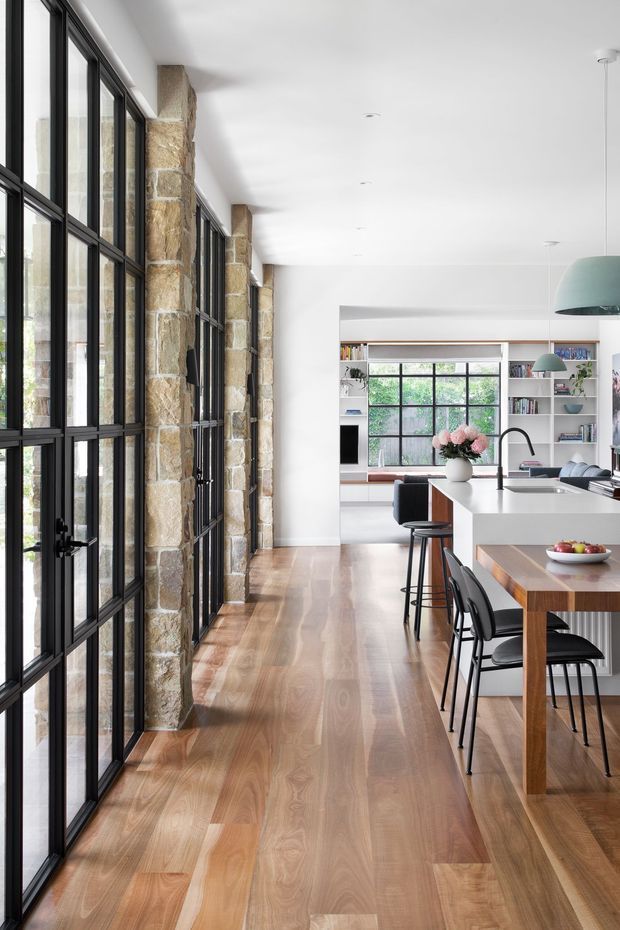 A bright, open-plan kitchen and dining area with extensive black-framed floor-to-ceiling glazing along one side, exposed rough stone piers, and warm timber flooring. The kitchen features a large white island with black bar stools, a timber dining table with black chairs, pale green pendant lights, and a built-in living area with shelving visible beyond.