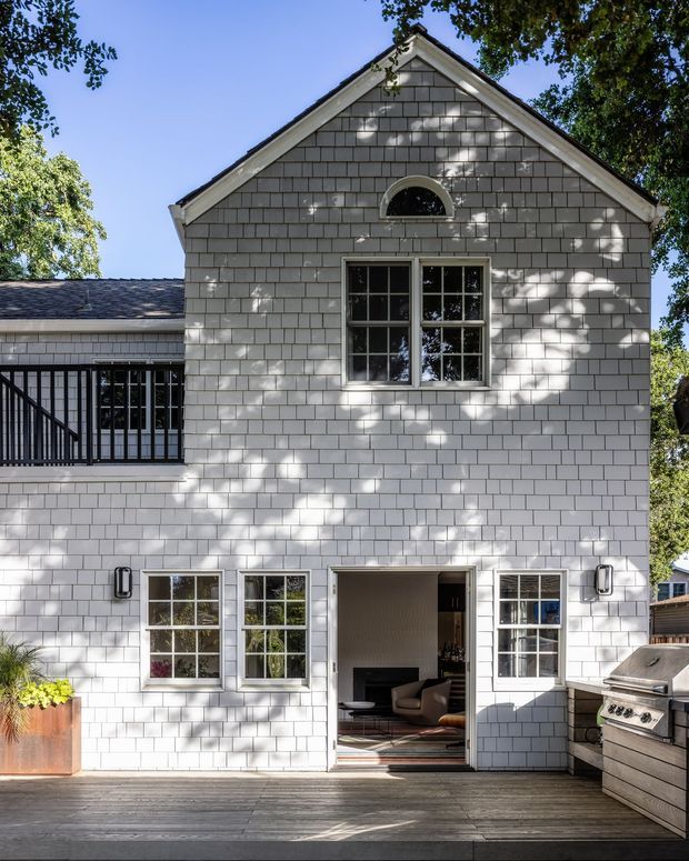 Exterior view of a light-coloured, shingle-clad two-storey house opening onto a timber deck. The symmetrical gabled facade has dark-trimmed multi-pane windows, a small arched upper window, an open ground-floor doorway, and a side balcony or enclosed outdoor connection. Mature trees cast dappled shadows across the facade, with an outdoor grill visible at the right.