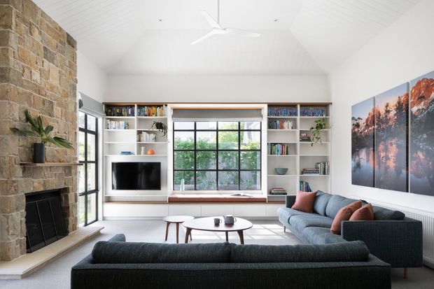Bright, spacious living room with a high vaulted white ceiling, large black-framed windows, built-in white shelving and cabinetry surrounding a television, and a substantial natural-stone fireplace wall. The room contains multiple dark upholstered sofas, a round coffee table, and a large panoramic landscape artwork. The overall design combines a clean contemporary fit-out with warm rustic stone detailing and abundant natural light.