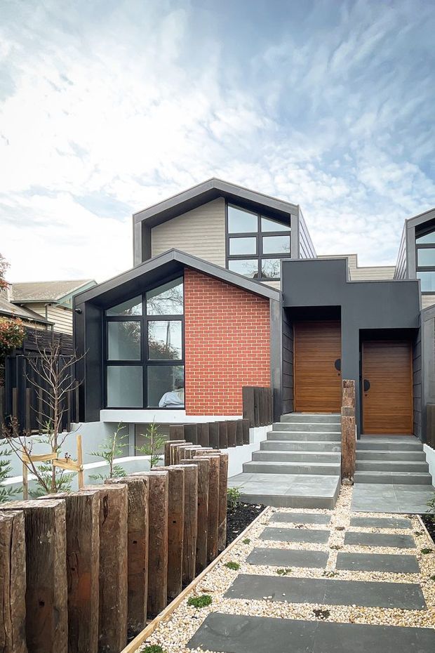 Street-facing view of a compact contemporary multi-level residence with a strong mix of red brick, dark painted cladding, timber-look doors, and extensive black-framed glazing. The home has intersecting pitched and skillion-like roof forms, a prominent glazed upper volume, concrete entry stairs, and a landscaped path edged with rustic timber posts.