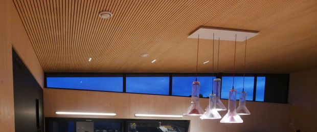 Interior architectural detail showing a warm timber slatted ceiling and matching timber wall lining, with recessed lighting, clerestory-style horizontal glazing, and a cluster of suspended glass pendant lights over a partially visible counter or workspace. The image has a refined contemporary fit-out with strong linear detailing and blue evening light visible through the upper windows.