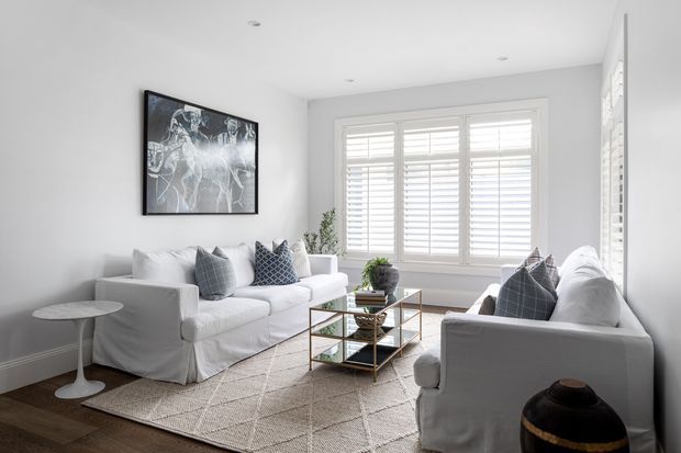 Bright, compact living room with two white upholstered sofas arranged around a glass-and-metal coffee table, layered over a light textured area rug. Large plantation-shuttered windows provide abundant natural light, while monochrome patterned cushions, a framed artwork, a small side table, and potted greenery add understated detail. The room has a clean, refined transitional feel with warm timber flooring and pale neutral walls.
