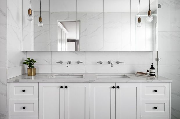 Bright, symmetrical contemporary bathroom vanity with extensive white cabinetry, twin integrated basins, wall-mounted chrome tapware, a large mirrored cabinet, and white marble-look tiled walls and benchtop. Pendant lights with brass fittings and clear glass shades add warm accents, while a small potted plant and toiletries provide restrained styling.