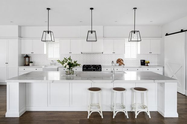 Bright, spacious white kitchen with extensive shaker-style cabinetry, a large marble-look island with seating for three, pendant lights, patterned backsplash, dark timber flooring, and a farmhouse-style door.