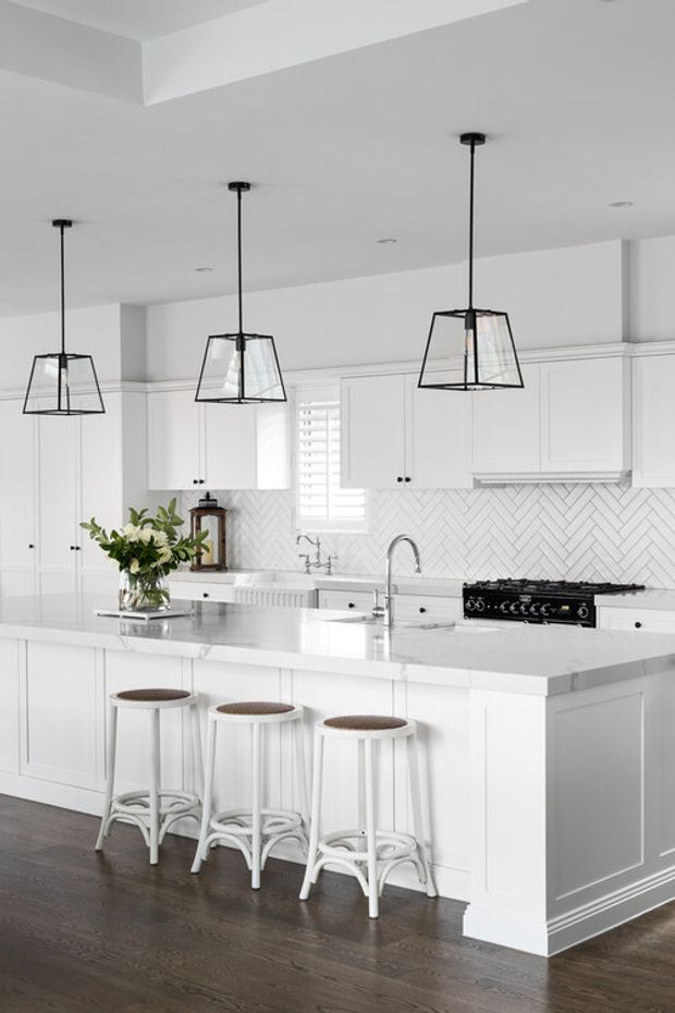Bright, spacious white kitchen with a large island, white shaker-style cabinetry, paneled island sides, white stone countertop, and a white herringbone tile backsplash. Three black geometric pendant lights hang above the island, which has three white stools with woven seats. Dark timber flooring provides contrast, while stainless-steel fixtures and a black range add subtle accents.