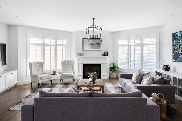 Bright, spacious living room with a large grey sectional sofa arranged around a wood-and-glass coffee table and a white fireplace mantel. Two tufted armchairs sit by the broad plantation-shuttered windows, while a geometric pendant light, framed artwork, built-in cabinetry, and indoor plants complete the refined neutral interior.