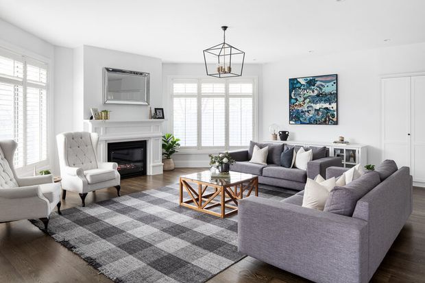 Bright, refined living room with white walls and plantation shutters, a white fireplace surround with dark insert, grey upholstered sofas and armchairs, a large grey-and-white checked rug, a geometric wood-and-metal coffee table, and a black geometric pendant light. Artwork and restrained accessories add colour while maintaining a clean, polished feel.