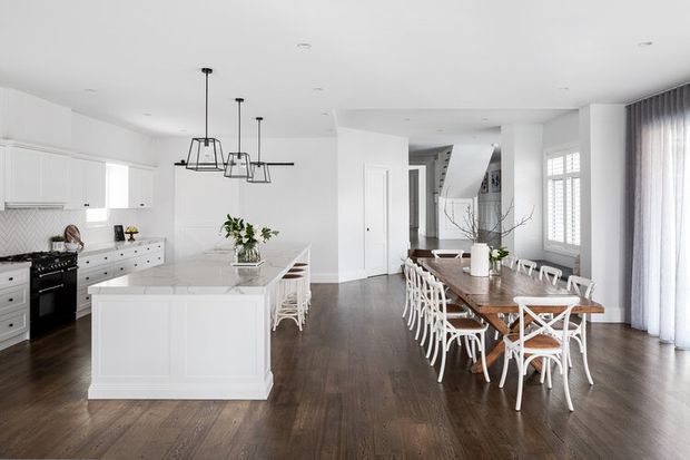 Bright open-plan kitchen and dining space with white shaker-style cabinetry, a large central island, dark timber flooring, and a long rustic timber dining table surrounded by white cross-back chairs. Large windows with sheer curtains provide abundant natural light, while simple black pendant lights add contrast. A hallway and staircase are visible beyond the dining area.