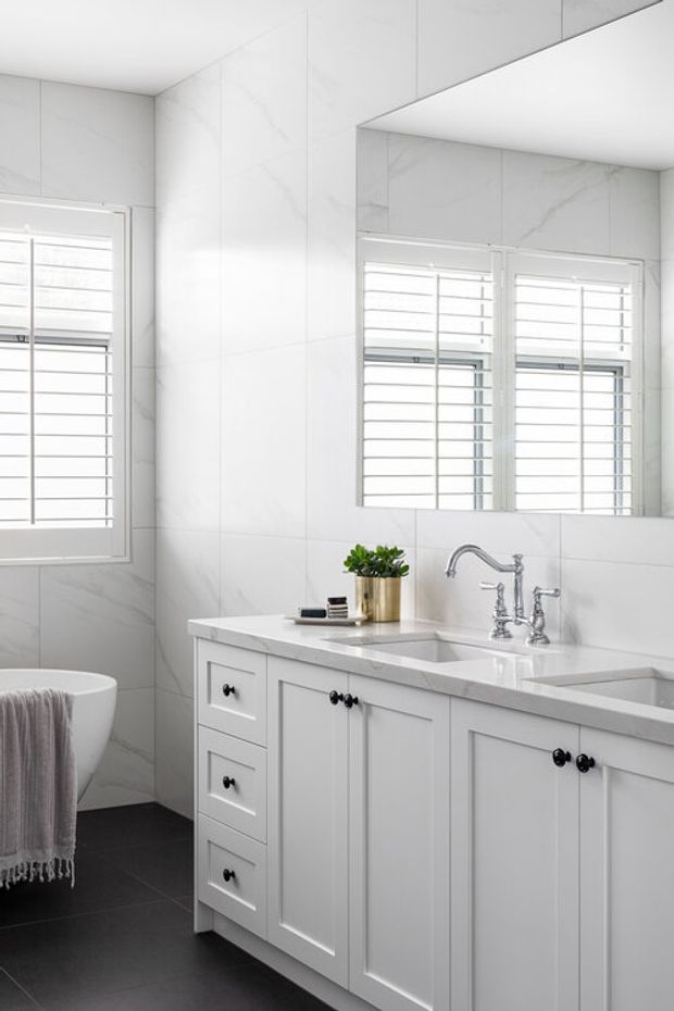 Bright, refined bathroom with white marble-look wall tiles, a large mirrored wall, plantation-shuttered windows, and a white traditional vanity with dark hardware. The vanity includes a pale stone benchtop, integrated basins, and polished silver tapware, contrasted by dark floor tiles and a partially visible freestanding bathtub.