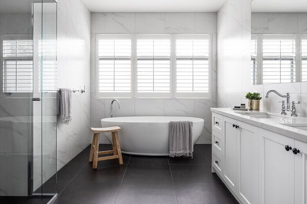 Bright, refined bathroom featuring a freestanding white bathtub beneath plantation-shuttered windows, a frameless glass shower enclosure at left, and a white vanity with marble-look benchtop and traditional hardware. Large dark floor tiles create contrast against the predominantly white walls and cabinetry.