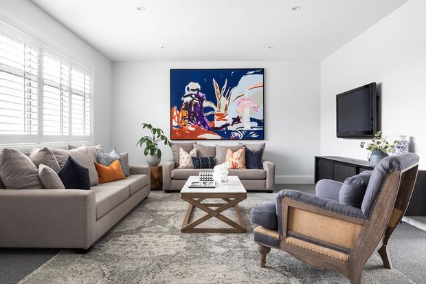 Bright, refined living room with neutral upholstered sofas and armchairs, layered cushions, a patterned area rug, a timber-framed coffee table, wall-mounted television, low dark media console, indoor plant, and colourful abstract artwork. White walls, recessed ceiling lights, and plantation shutters create a clean contemporary setting.