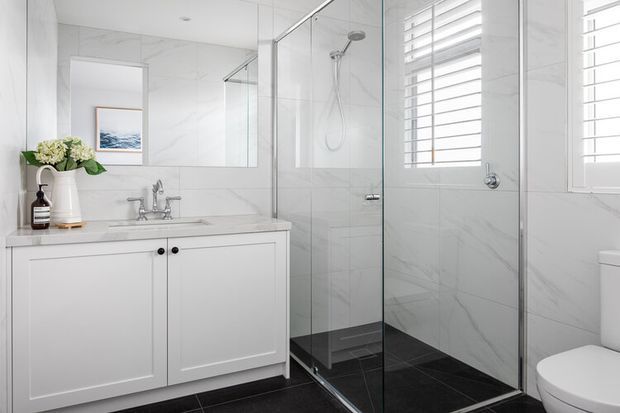 Bright, refined bathroom with white marble-look wall and vanity surfaces, a white panelled vanity with chrome tapware, frameless glass shower enclosure, dark tiled shower floor, plantation shutters, and a partially visible toilet. A large mirror enhances the light and spacious feel.