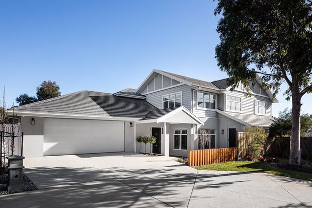 A large two-storey suburban residence with a light grey weatherboard-style exterior, white trim, multiple gabled roof sections, and an attached double garage. The front entry has a small projecting gabled porch, with a concrete driveway, lawn, mature trees, and timber fencing visible around the property.