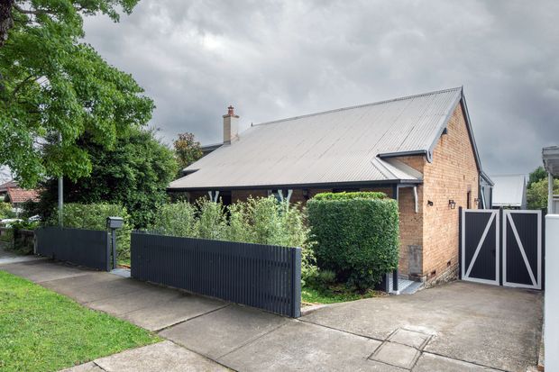 Street-facing exterior view of a single-storey brick house with a steeply pitched light metal gable roof, chimney, dark metal fencing and gates, hedges, and a concrete driveway and footpath. The property has a simple suburban form with landscaped frontage under an overcast sky.