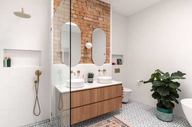 Bright contemporary bathroom with white wall tiles, a warm timber floating double vanity, arched mirrors, exposed brick feature wall, brass fixtures, patterned encaustic-style floor tiles, and a glass shower enclosure. A freestanding bathtub and large indoor plant add softness to the space.
