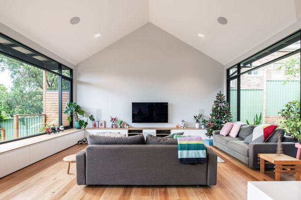Bright contemporary living room with a high pitched ceiling and expansive black-framed corner glazing. A large grey sectional sofa faces a wall-mounted television, with built-in low cabinetry along the window wall, timber flooring, indoor plants, and Christmas decorations. The glazing overlooks a leafy suburban setting and deck or balcony area.