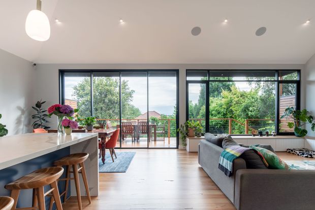 Open-plan contemporary living space combining a kitchen island with bar stools, dining area, and living room. Large black-framed glazed doors and windows open to a timber deck and leafy outdoor setting. The room has pale timber flooring, white walls and ceiling, a light benchtop, recessed lighting, indoor plants, and colourful soft furnishings.