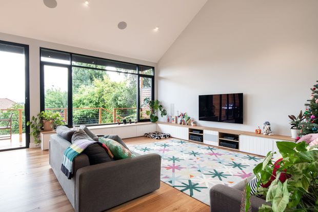 A bright, contemporary living room with large black-framed windows and glazed doors opening toward a leafy outdoor setting. The room features a grey sectional sofa, timber flooring, a large colourful geometric rug, a wall-mounted television, and extensive white built-in media cabinetry. Indoor plants and a Christmas tree add decorative detail.