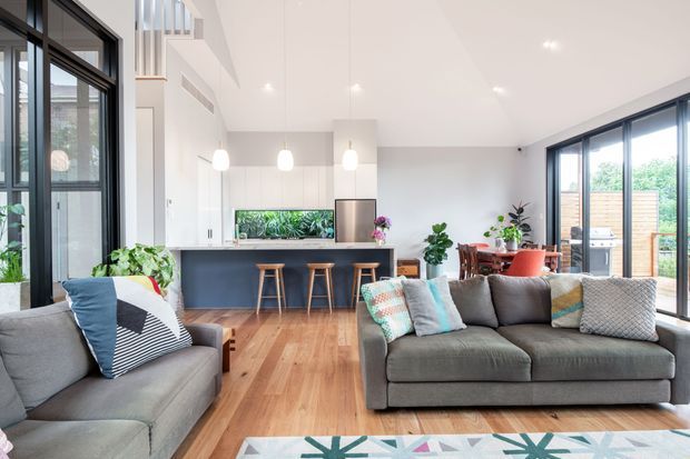 Bright open-plan contemporary living area with a modern kitchen at the rear, a large island with timber stools, grey sofas, indoor plants, and a dining area beside extensive floor-to-ceiling glazing. Warm timber flooring contrasts with white walls and dark-framed windows and doors. A partial upper-level void or stair landing is visible, creating an airy, spacious feel.