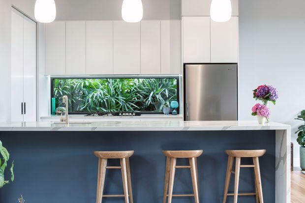 Bright contemporary kitchen with white full-height cabinetry, a large white island with a dark navy feature front, three timber bar stools, stainless-steel refrigerator, and pendant lights. A wide window above the sink looks onto dense greenery, while flowers and indoor plants add colour. The room has a clean, fresh, high-contrast feel.