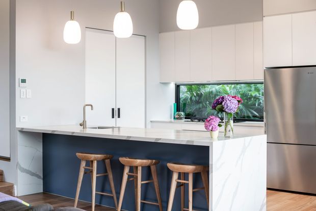 Bright contemporary kitchen with a large white stone island and integrated sink, dark blue island cabinetry, three timber bar stools, white overhead cabinetry, pendant lights, and a stainless-steel refrigerator. A horizontal window above the benchtop overlooks lush greenery, while pale timber flooring and minimal detailing create a clean, airy interior.