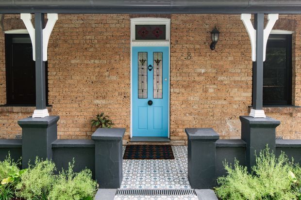 Front entry facade of a traditional brick residence, featuring a vivid turquoise panelled front door with decorative glazed inserts, black-framed windows, dark-painted porch columns and low rendered masonry walls. A patterned tiled pathway leads to the entrance, with planting along both sides.