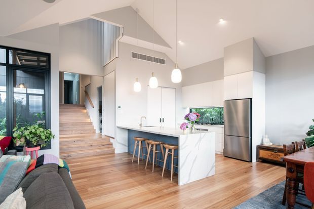 Bright open-plan kitchen and living area with a contemporary white kitchen, central island with timber stools, stainless-steel refrigerator, white marble-look island panels, timber flooring, and a timber staircase leading to an upper level. The room has a high sloping ceiling, pendant lights, large black-framed glazing, indoor plants, and a partially visible living and dining area.