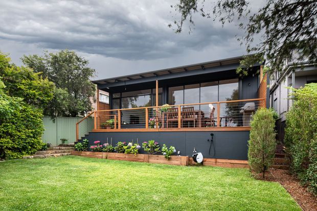 A contemporary single-storey backyard extension or pavilion with dark cladding, extensive glazed doors, and a covered timber-framed deck. The deck includes outdoor dining furniture and a barbecue, with raised garden beds and a neatly maintained lawn in the foreground. The setting is leafy and suburban beneath an overcast sky.