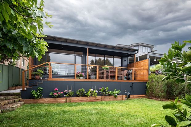 Exterior view of a contemporary residential extension with a covered glazed outdoor living area, dark cladding, warm timber decking and horizontal timber privacy screens. The elevated terrace has wire balustrading, outdoor furniture and potted plants, opening onto a landscaped lawn and garden. The home has a low-pitched roof and large floor-to-ceiling glazing beneath an overcast sky.