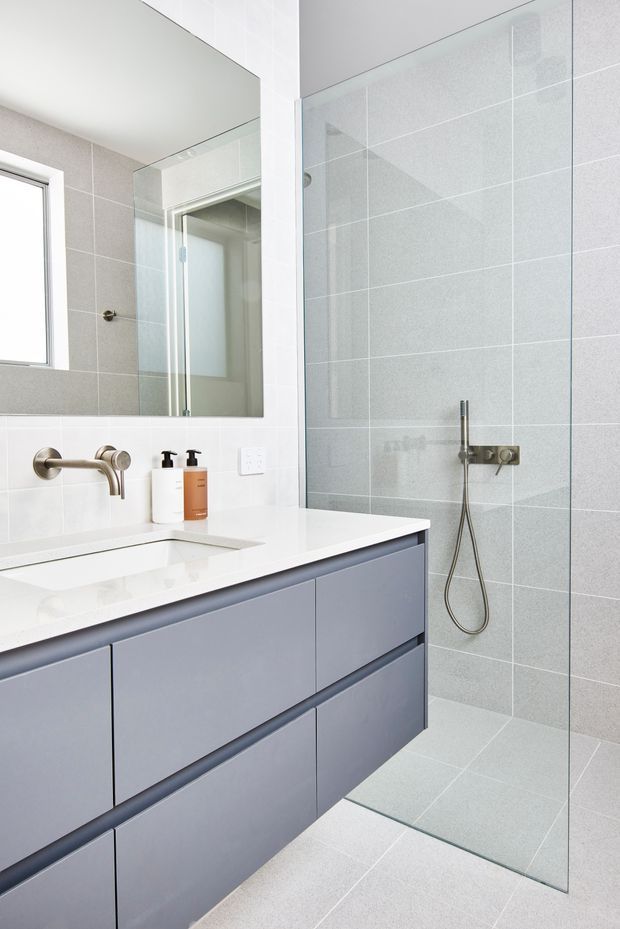 Bright contemporary bathroom with a frameless glass walk-in shower, pale grey tiled walls and floor, a floating blue-grey vanity with a white stone benchtop, wall-mounted brushed metal tapware, and a large mirror.