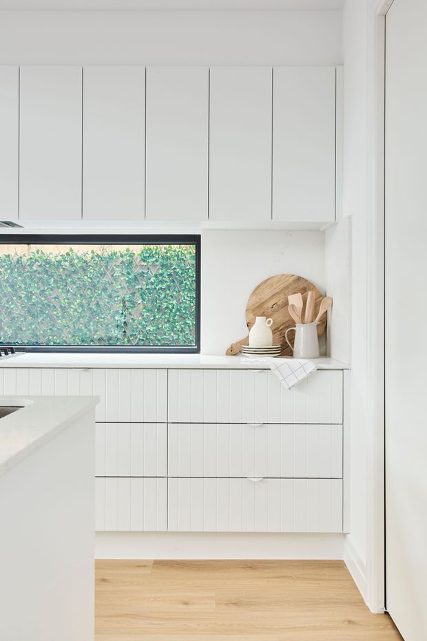 Bright, compact white kitchen with extensive vertical-panel cabinetry, a white benchtop, black-framed horizontal window, open shelf styling, and pale timber flooring. The cabinetry and walls create a clean, understated contemporary look with natural wood accessories and greenery visible outside the window.