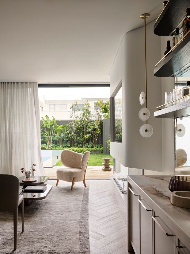 A bright, refined open-plan living and dining area with a contemporary built-in bar or kitchen joinery. The room features pale walls, full-height curtains, a large glazed opening to a landscaped courtyard, a sculptural upholstered armchair, dark dining furniture, a low coffee table, and a light rug. White cabinetry with dark linear handles and a stone benchtop occupies the foreground, while open dark timber shelves display glassware and bottles. Light timber herringbone flooring leads toward the outdoor deck and garden.
